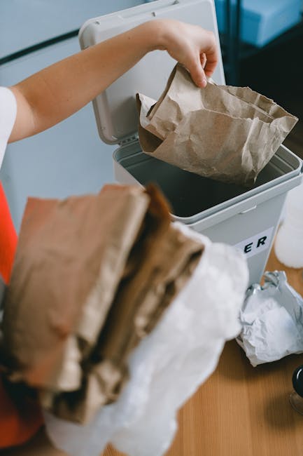 A person is disposing of crumpled brown and white paper waste into a white plastic bag or container, which is partially open and positioned on a wooden surface. The person's hand is gripping the paper by the edges, and they are wearing a short-sleeved shirt, with a portion of their forearm visible. The background shows a white bin or designated waste container with the word 'RUBBISH' printed on it in black. Additional crumpled paper is seen nearby on the surface, indicating the ongoing tidying or waste removal process. The environment appears to be indoors, with a modern and tidy setting, suitable for waste collection activities. The scene reflects general waste handling practices, consistent with rubbish removal services like those provided by Waste Collection Enfield, without depicting specific waste types or collection equipment.