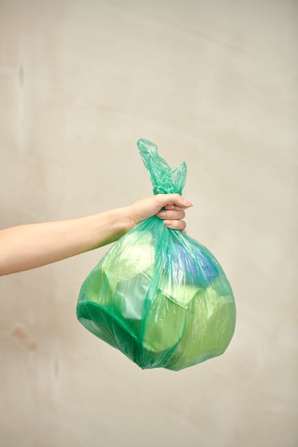 A person is disposing of crumpled brown and white paper waste into a white plastic bag or container, which is partially open and positioned on a wooden surface. The person's hand is gripping the paper by the edges, and they are wearing a short-sleeved shirt, with a portion of their forearm visible. The background shows a white bin or designated waste container with the word 'RUBBISH' printed on it in black. Additional crumpled paper is seen nearby on the surface, indicating the ongoing tidying or waste removal process. The environment appears to be indoors, with a modern and tidy setting, suitable for waste collection activities. The scene reflects general waste handling practices, consistent with rubbish removal services like those provided by Waste Collection Enfield, without depicting specific waste types or collection equipment.