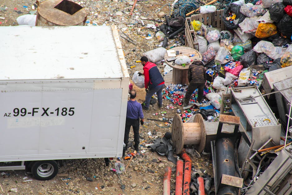 An aerial view of an outdoor waste disposal site featuring a large white container with the label 'AZ 99-FX-185' on its side, situated on rocky ground. Surrounding the container is a significant accumulation of mixed waste including plastic bags, crumpled packaging, and discarded household items. Several people are actively engaged in sorting or collecting rubbish; one person is wearing a red and black jacket, another in dark clothing, and a third in a purple top, all focused on handling and managing the waste. Nearby, there are wooden pallets, large spools of cable or piping, and various metal and plastic objects, including a washing machine and other appliances. The environment appears cluttered, with waste spilling from the containers and scattered across the uneven ground, highlighting the context of private waste management or alternative rubbish collection solutions, as operated by Waste Collection Enfield. Natural light illuminates the scene, emphasizing the textures and colors of the waste materials, which range from bright plastic to dull metallic surfaces.