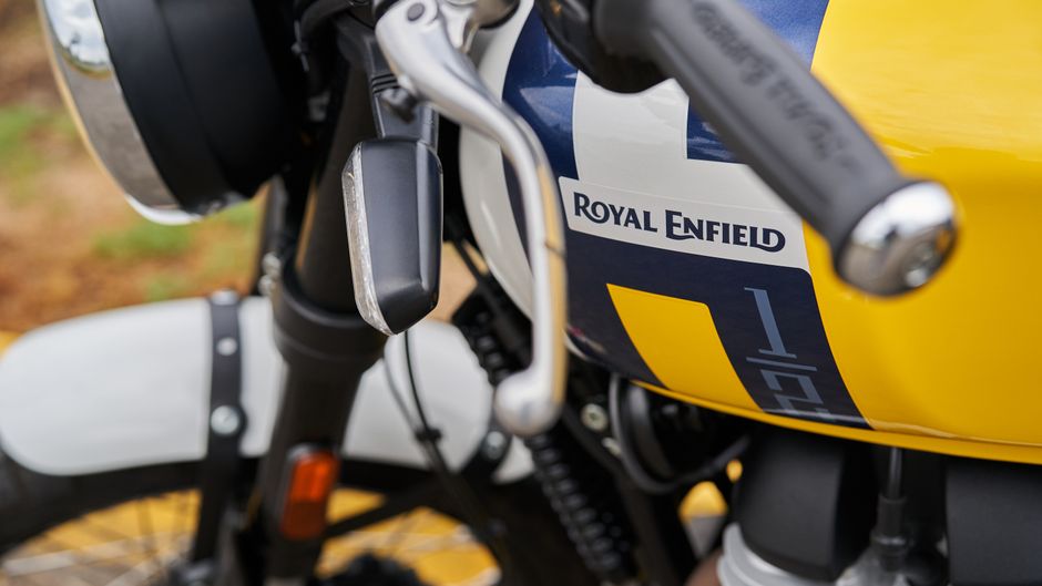 Close-up image of a Royal Enfield motorcycle showing the front section, including the handlebar, speedometer, and part of the fuel tank. The fuel tank is painted in yellow with a black and blue Royal Enfield logo and a numbering decal. The handlebar features a metallic brake lever and the left side component, with textured grips visible. The front fork is black, with a white front fender curving over the wheel, which has a yellow rim matching the fuel tank. The background is blurred, revealing an outdoor setting with a mix of earthy ground and greenery, consistent with a parking area or open space. The image highlights the motorcycle's clean, polished surface and detailed textures, aligning with the context of transport or vehicle services related to rubbish removal or private waste transportation from a collection perspective, with subtle visual cues of vehicle management and alternative waste handling options.