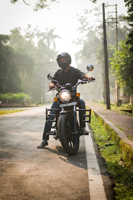 A person wearing a black helmet, black face covering, and black clothing is seated on a black motorcycle parked on the edge of a paved street. The motorcycle's front headlight is illuminated and it has a sturdy metal front rack and side cases for storage. The street is lined with a narrow sidewalk and grassy verge, with tall trees and utility poles visible in the background, suggesting a suburban or rural environment. The scene appears to be set in the early morning or late afternoon due to the soft, diffused lighting and slight haze in the atmosphere. The setting offers a neutral view of roadside urban or suburban waste management, with no visible rubbish or debris. The image’s focus on the motorcycle and rider, along with natural surroundings, aligns subtly with themes of local transportation and the importance of safe, personal mobility, possibly supporting a message about alternative waste removal methods like on-site collection or independent disposal options provided by waste services such as Waste Collection Enfield.
