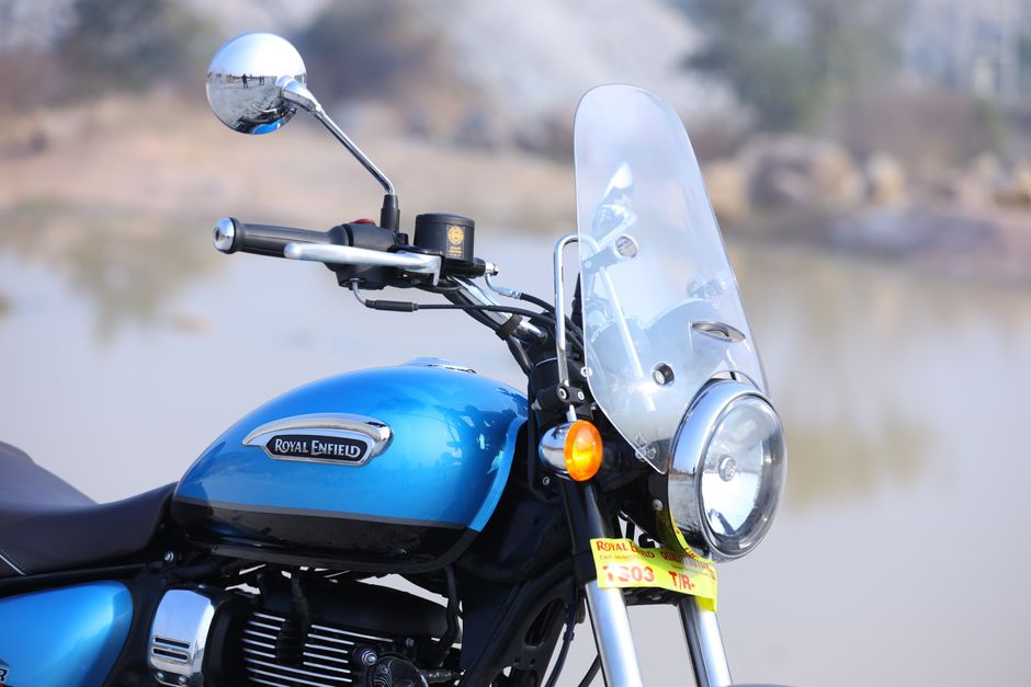 Close-up image of a Royal Enfield motorcycle showing the front section, including the handlebar, speedometer, and part of the fuel tank. The fuel tank is painted in yellow with a black and blue Royal Enfield logo and a numbering decal. The handlebar features a metallic brake lever and the left side component, with textured grips visible. The front fork is black, with a white front fender curving over the wheel, which has a yellow rim matching the fuel tank. The background is blurred, revealing an outdoor setting with a mix of earthy ground and greenery, consistent with a parking area or open space. The image highlights the motorcycle's clean, polished surface and detailed textures, aligning with the context of transport or vehicle services related to rubbish removal or private waste transportation from a collection perspective, with subtle visual cues of vehicle management and alternative waste handling options.