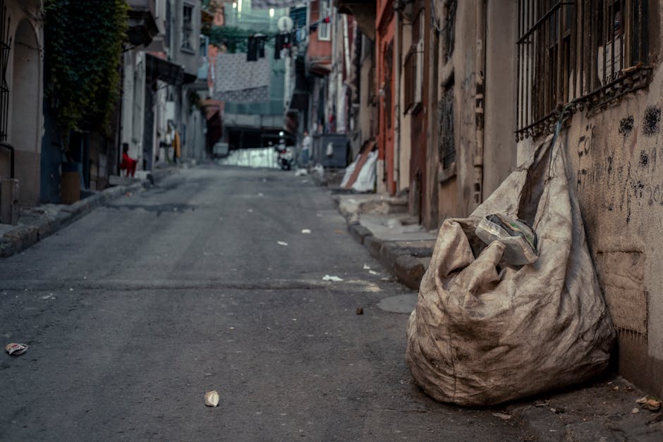 A large, worn beige trash bag made of heavy-duty plastic is placed on the pavement beside a weathered brick and concrete building. The bag appears full, with some items visible at the top, including a transparent plastic container and miscellaneous small debris. The bag is situated near a black metal security grille covering a window, with graffiti marks on the wall behind it. The street scene extends into a narrow alleyway with uneven, slightly sloped asphalt providing a textured surface with scattered litter. Distant residential buildings with laundry hanging on clotheslines are visible further along the street, which is lined with varied aged facades, some with peeling paint and exposed brickwork. Natural lighting suggests daytime, with soft shadows cast on the ground. The scene conveys an environment where private or independent waste disposal near residential areas, such as through rubbish collection services like Waste Collection Enfield, might be necessary to manage urban cleanliness and waste handling effectively.