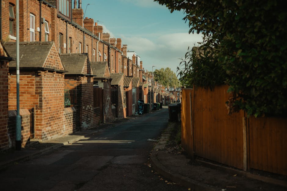A narrow residential street lined with terraced brick houses featuring pitched roofs, with small front gardens enclosed by brick walls and a sidewalk on the left side. On the right, a tall wooden fence runs alongside dense green foliage, partially obscuring the view. The street surface is asphalt, with some uneven patches and scattered leaves. Several black wheelie bins, used for rubbish collection, are positioned along the sidewalk and near the houses, indicating a typical urban waste management setting. The scene is illuminated by natural daylight, with soft shadows cast on the ground, suggesting late afternoon or early evening. The environment reflects a quiet neighbourhood where private waste disposal and on-site rubbish collection services, such as those offered by Waste Collection Enfield, are commonly used to manage refuse on narrow streets where vehicle access may be limited. The overall atmosphere is calm, clean, and organized, with no visible litter or debris on the street surface.