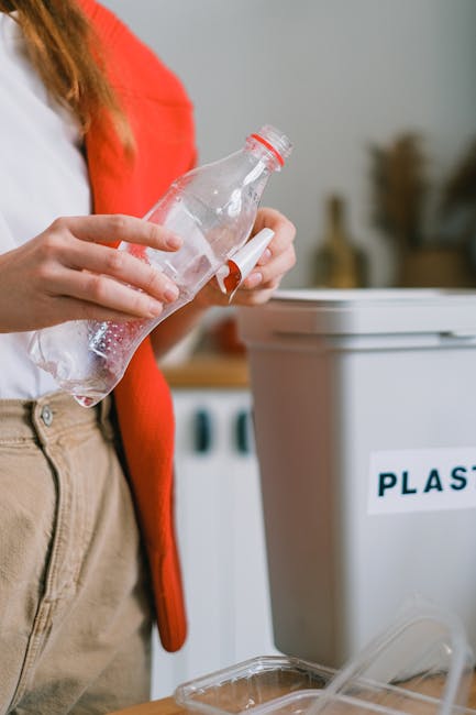 A person wearing beige trousers and a white shirt with a red cardigan is holding a clear plastic bottle over a recycling bin labeled 'PLASTIC'. The individual is in the process of preparing the bottle for disposal or recycling, with a focus on responsible waste sorting. The background includes a blurred view of a domestic kitchen setting, with visible appliances and indoor elements. Nearby, there is a transparent plastic container, possibly for separating recyclables or waste, positioned on a wooden surface. The scene emphasizes proper waste management and the importance of separating plastic items for recycling, which aligns with domestic rubbish removal and environmentally conscious waste handling services provided by Waste Collection Enfield. The lighting appears natural, contributing to a neutral, professional atmosphere suitable for an informational or service-related context about alternative rubbish collection methods such as private recycling or on-site waste disposal. The attention to detail in the act of recycling reinforces themes of environmentally responsible waste management and the role of independent collection services in managing household rubbish.