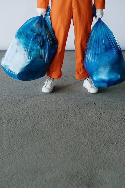 A person standing on a grey indoor floor holding two large blue rubbish bags, one in each hand, with the bags appearing to be full of waste materials. The individual is dressed in orange work overalls and white sneakers, with the lower half of their body visible from the ankles to the waist. The background shows a plain, light-colored wall, suggesting an indoor environment such as a warehouse or storage area where waste collection or disposal activities are carried out. The scene illustrates private waste handling or on-site clearance, typical of waste removal services, with the emphasis on the bags' size and the professional clothing indicating a waste collection context, consistent with services offered by Waste Collection Enfield or similar providers for rubbish removal and bulky item disposal.