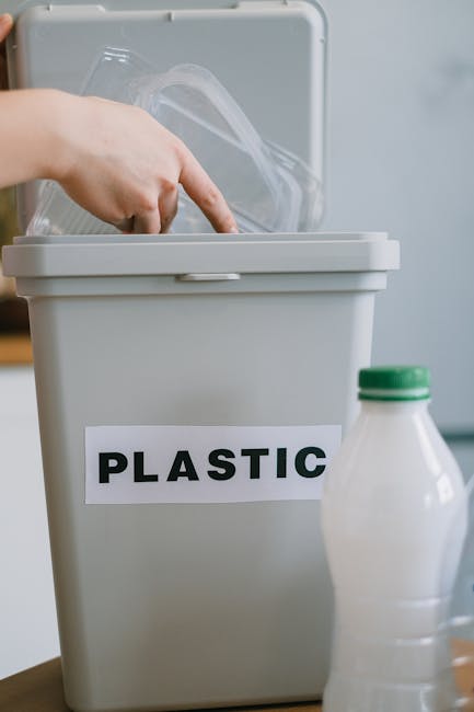 A person wearing beige trousers and a white shirt with a red cardigan is holding a clear plastic bottle over a recycling bin labeled 'PLASTIC'. The individual is in the process of preparing the bottle for disposal or recycling, with a focus on responsible waste sorting. The background includes a blurred view of a domestic kitchen setting, with visible appliances and indoor elements. Nearby, there is a transparent plastic container, possibly for separating recyclables or waste, positioned on a wooden surface. The scene emphasizes proper waste management and the importance of separating plastic items for recycling, which aligns with domestic rubbish removal and environmentally conscious waste handling services provided by Waste Collection Enfield. The lighting appears natural, contributing to a neutral, professional atmosphere suitable for an informational or service-related context about alternative rubbish collection methods such as private recycling or on-site waste disposal. The attention to detail in the act of recycling reinforces themes of environmentally responsible waste management and the role of independent collection services in managing household rubbish.