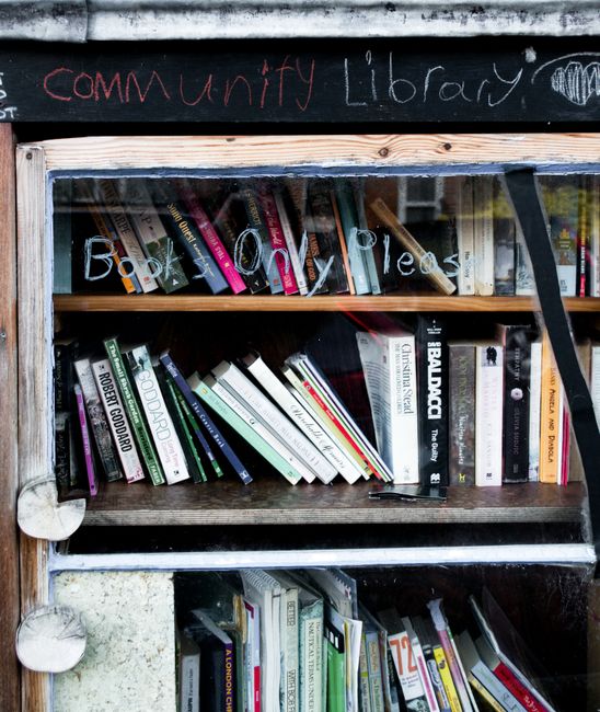 A wooden bookshelf situated outdoors, displaying a collection of books arranged in multiple horizontal shelves. The top shelf contains books with various colored covers, some leaning against the sides while others stand upright, with titles that include fiction and non-fiction genres. The middle shelf features more books of different sizes, stacked and leaning at angles, with visible titles on their spines. The bottom shelf shows additional books, some of which extend beyond the edge, with a noticeable mix of hardcover and paperback editions. Above the bookshelf, a black chalkboard with handwritten text in red and white chalk reads 'community Library,' indicating an informal sharing space for reading materials. The surroundings suggest an outdoor environment, possibly a garden or community space, with natural light illuminating the scene and slight weathering on the wood surfaces. This setup could be part of an initiative to encourage book sharing or promote resource exchange, aligning with community-driven waste reduction and reuse efforts facilitated by local waste management organizations like Waste Collection Enfield.