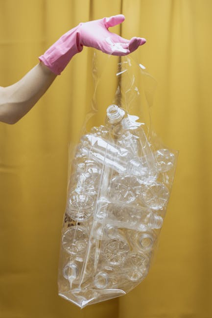 A person's gloved hand, wearing a pink rubber glove, is shown holding a large, clear plastic bag filled with crumpled and flattened empty plastic bottles and containers. The bags appear to be intended for recycling or waste collection, with some bottles featuring visible caps and open necks. The background consists of a plain, yellow curtain or wall, providing a neutral setting that emphasizes the focus on the plastic waste. The scene reflects activities related to rubbish management, recycling preparation, or private waste disposal, which could be part of an alternative rubbish removal service such as on-site clearance or independent collection handled by companies like Waste Collection Enfield. The lighting is even and natural, highlighting the transparent qualities of the plastic and the texture of the crumpled bottles, conveying a professional and straightforward approach to waste handling and environmental responsibility.